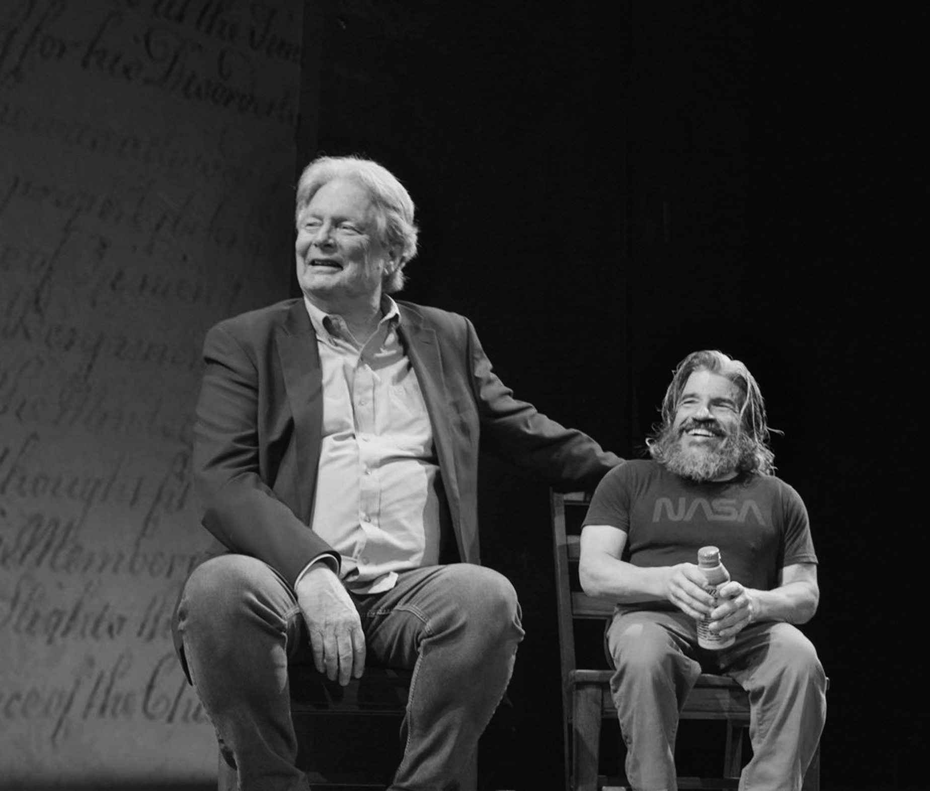 Black and white photo of Marcus Rediker and Mark Povinelli sitting on a stage in chairs, smiling towards the camera.